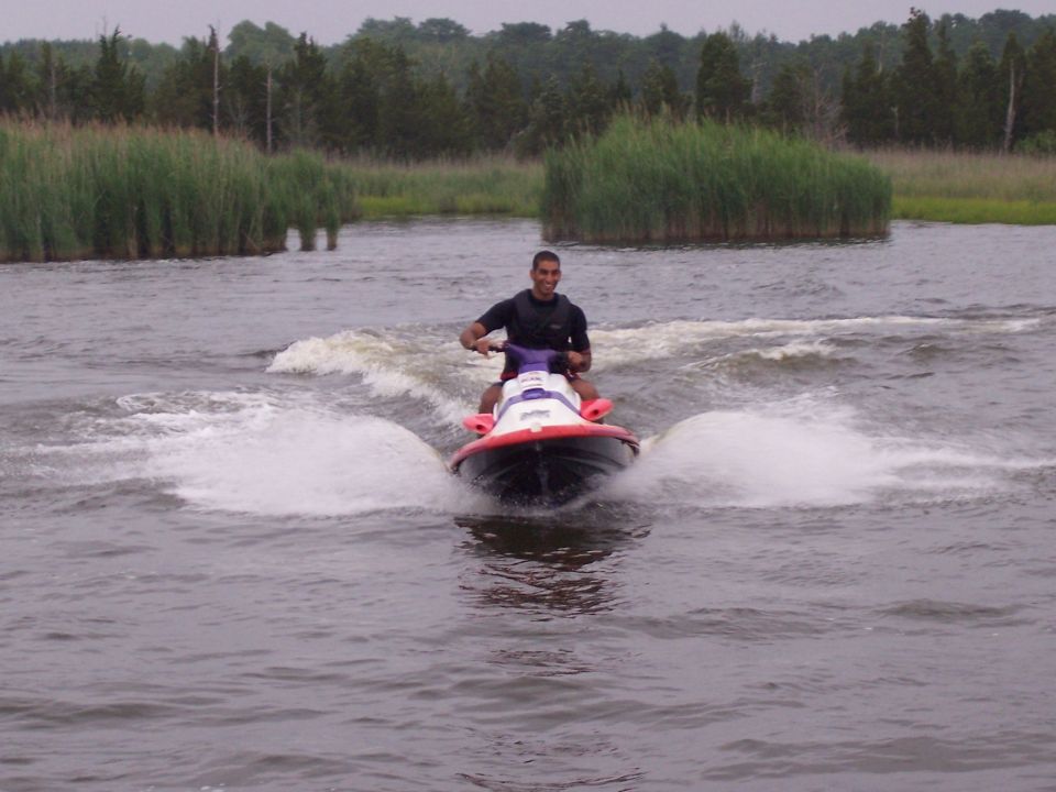 Man riding red and white jet ski toward camera on calm water with marsh in background