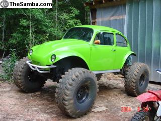 1990 Ford Bronco - bright green VW Beetle-bodied monster truck sits on massive tires and a tall lift beside a wooden shed.