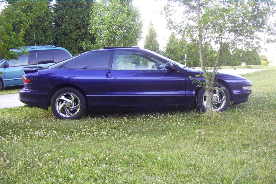 Purple 1996 Ford Probe parked on grass next to a tree. A blue minivan is visible in the background