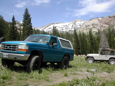 1994 Ford Bronco - Two off-road vehicles are parked in a scenic mountain landscape under a blue sky with scattered clouds.