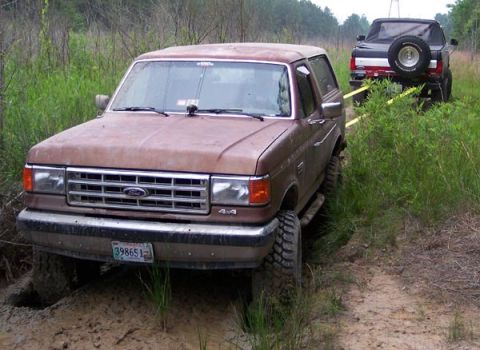 1988 Ford Bronco - A brown 1987 Ford Bronco is stuck in the mud while a black truck tows it with yellow straps.