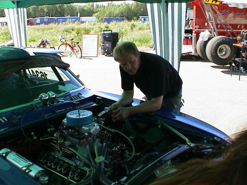 1979 Ford Bronco - Man works on the engine of a blue car, with racing equipment in the background.