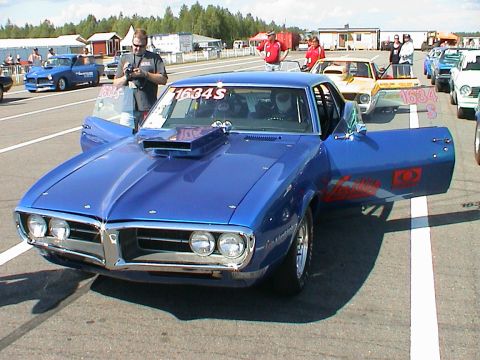 1979 Ford Bronco - A blue muscle car with racing decals sits on the track, doors open, ready for the next event.