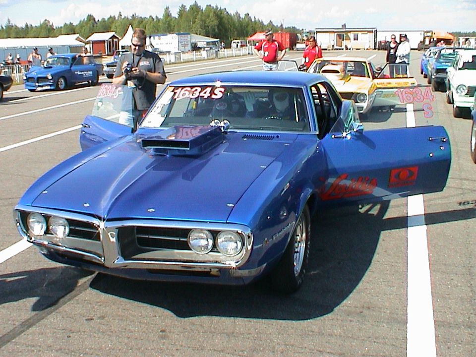 Blue drag car with hood scoop and open doors, parked on a track with other cars and people around