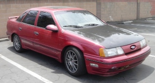 1991 Ford Taurus - Small three-quarter front photo of a red first-generation Ford Taurus SHO sedan parked on asphalt in front of a beige block wall. The car h…