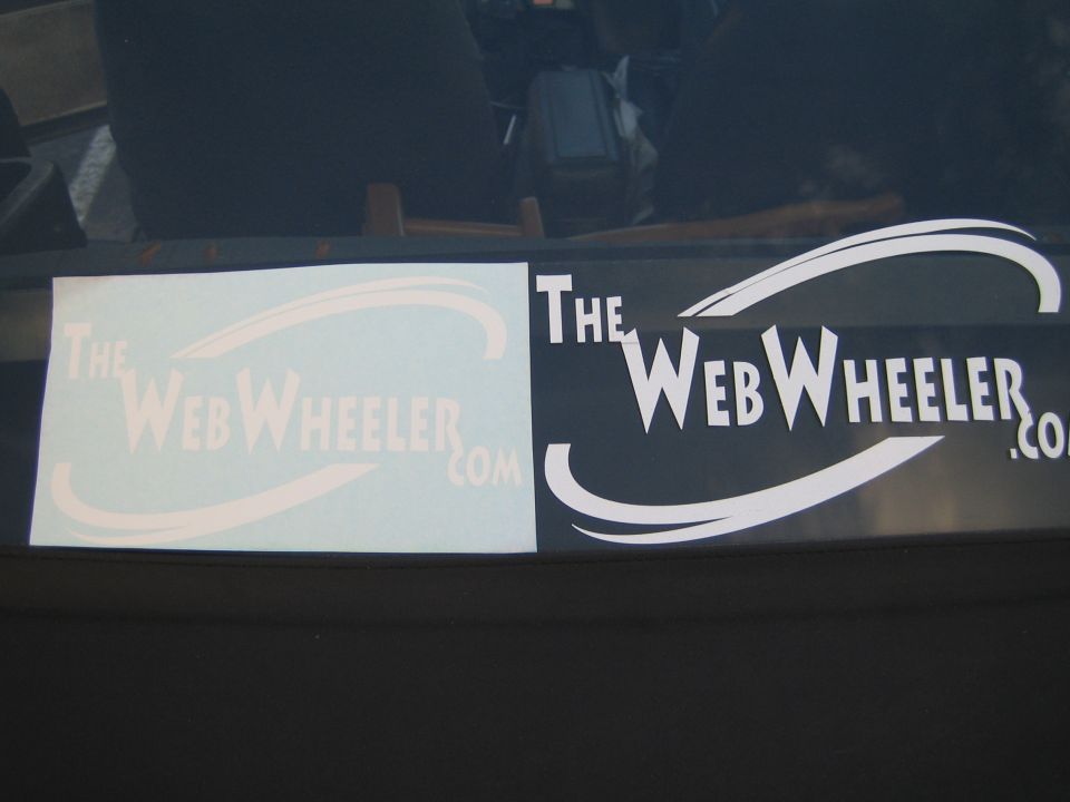 Two TheWebWheeler.com vinyl decals, one white and one printed, on a dark vehicle window