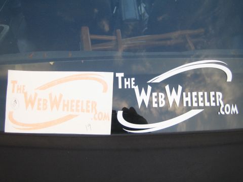 1990 Ford Bronco - Orange and white 'TheWebWheeler.com' decals on a car's rear glass reflecting a cloudy sky.