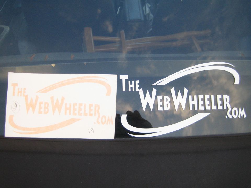 Orange and white 'TheWebWheeler.com' decals on rear window with sky reflection