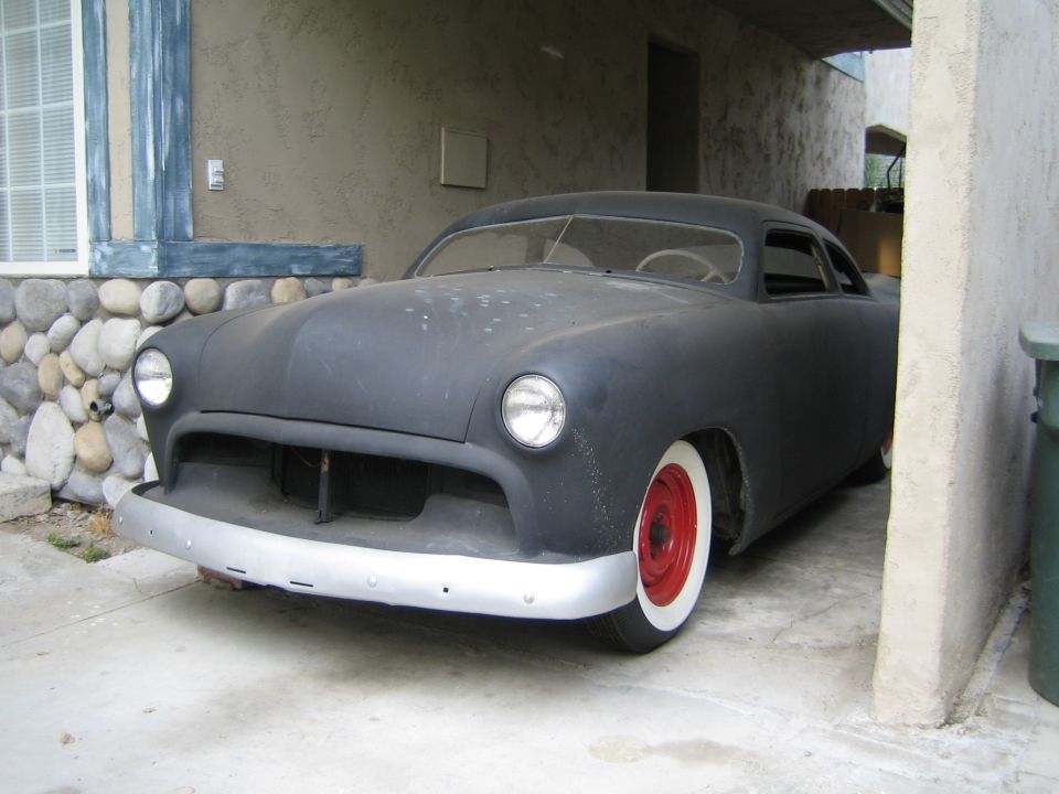 Front view of a matte black customized coupe hot rod with red wheels and whitewall tires parked under an overhang