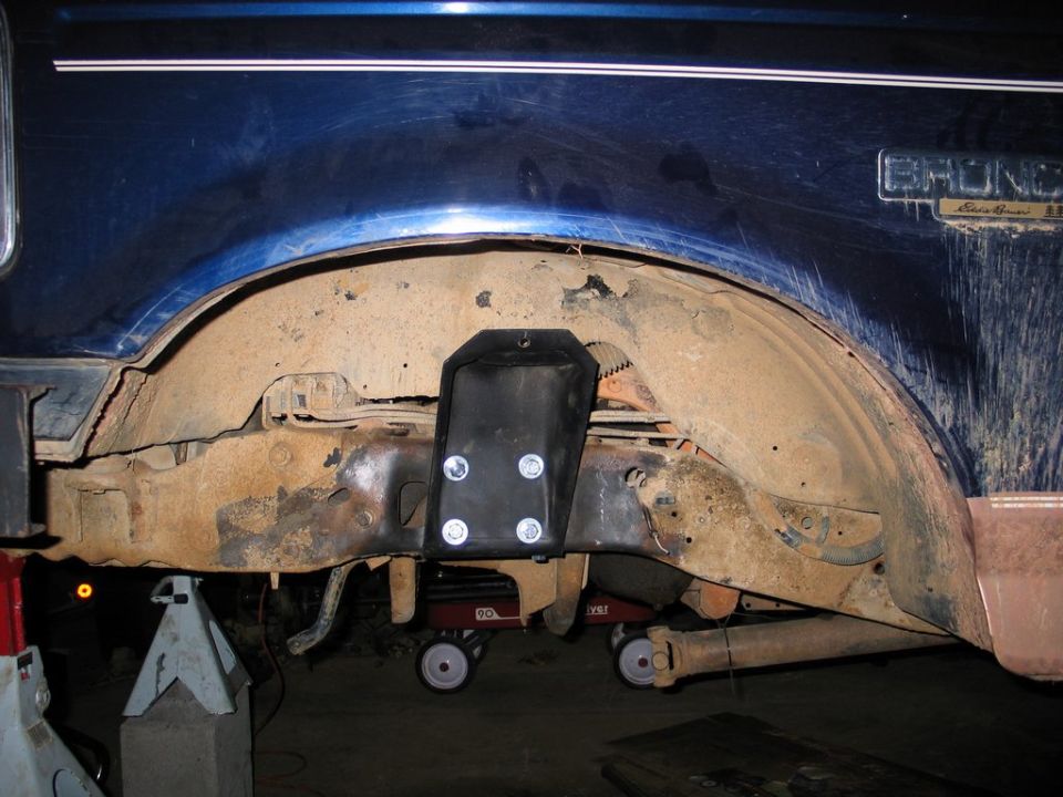 Underside of blue Ford Bronco on jack stands showing frame, wheel well, and bolted bracket