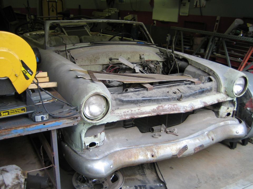 car body in workshop, hood open, engine exposed, front end disassembled, yellow chop saw nearby