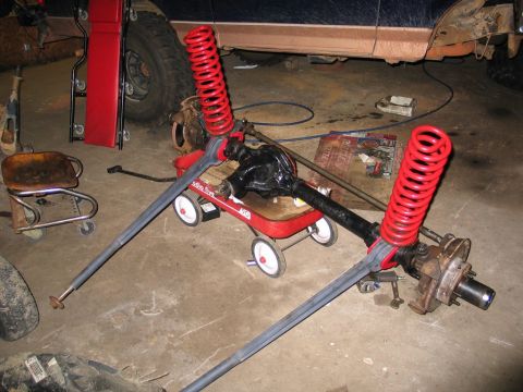 1995 Ford Bronco - A front axle assembly with red coil springs is placed on a red wagon, likely for a vehicle modification project.