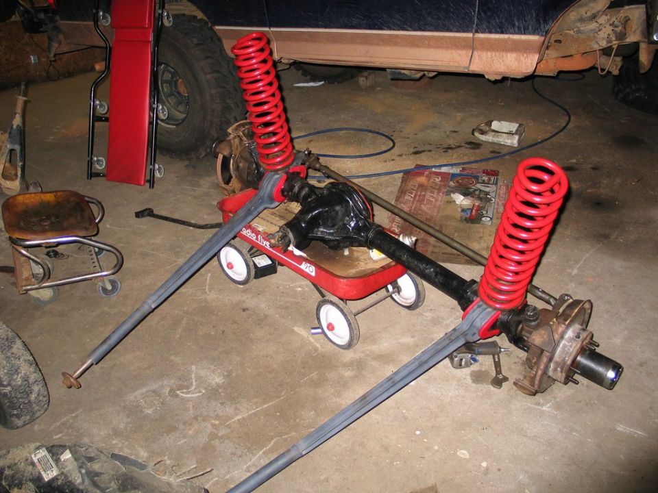 Black solid front axle with red coil springs and radius arms assembled on a Radio Flyer wagon in a garage