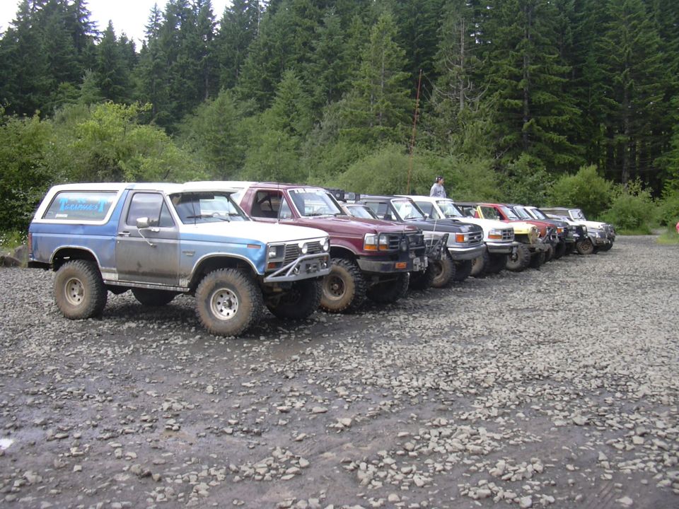 Line of modified Ford Broncos parked on gravel lot; front Bronco is blue and white with custom bumper