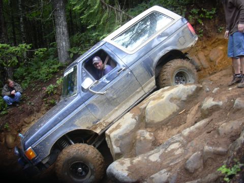 1991 Ford Bronco - A blue Ford Bronco is shown attempting to climb over large rocks on a muddy, wooded trail.