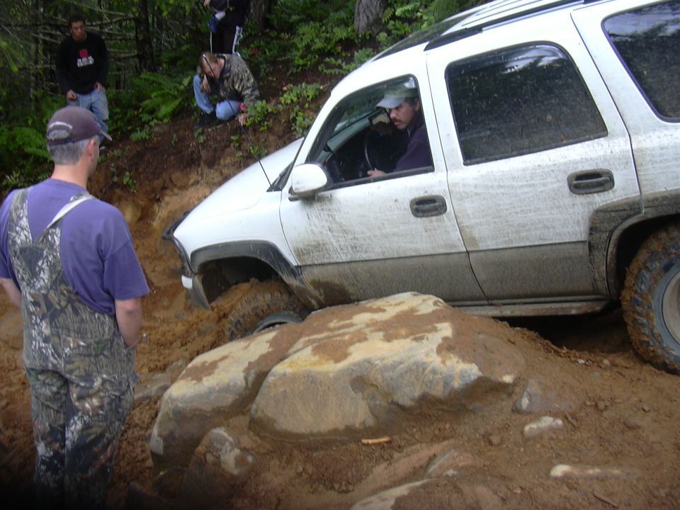 White SUV with driver visible, stuck on a muddy, rocky trail with people watching