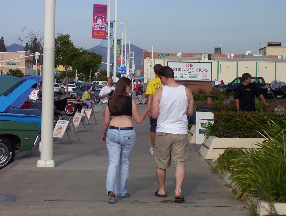 People walk past parked cars at an outdoor event, with buildings and mountains in the background