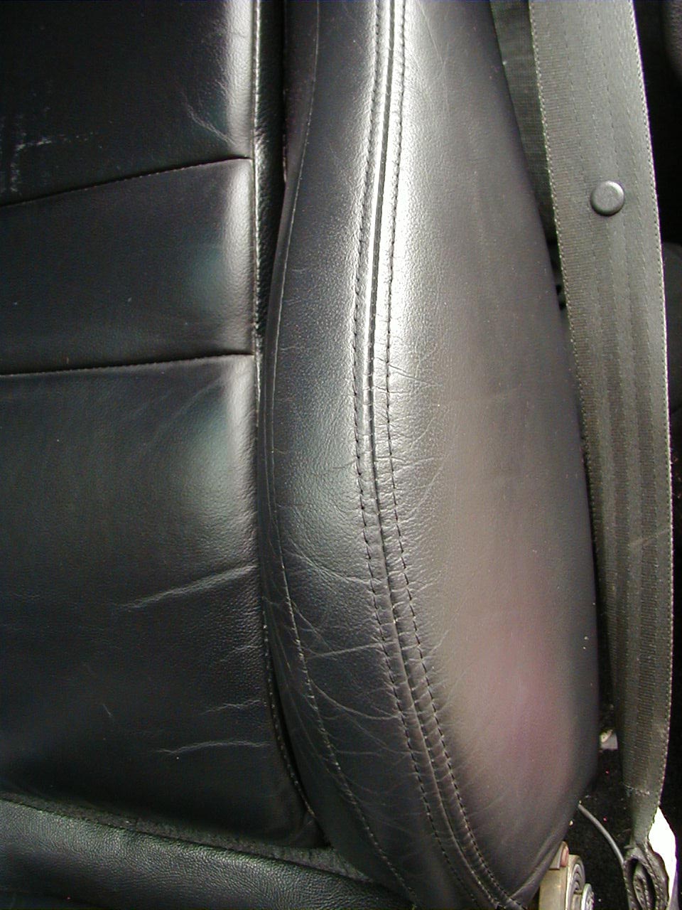 Close-up of black leather car seat with visible stitching and a seatbelt. Supermotors.net logo in corner