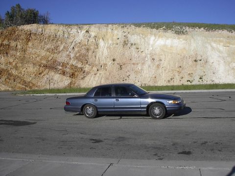 1985 Mercury Grand Marquis - A blue sedan is parked on a paved surface with a large dirt and grass embankment in the background under a clear blue sky.