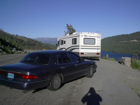 1985 Mercury Grand Marquis - A dark sedan is parked on the side of a road with an RV behind it, with a scenic view of a lake and mountains.