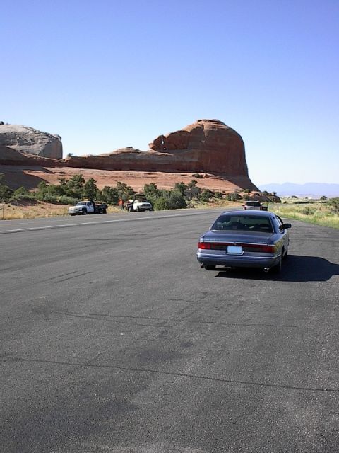 1985 Mercury Grand Marquis - Sedan on a road with rock formations and two trucks in the distance.