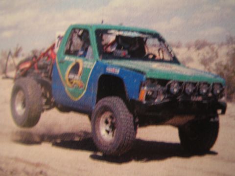 1990 Ford Bronco - green and blue desert race truck with multiple light pods catches air on sandy off-road terrain.