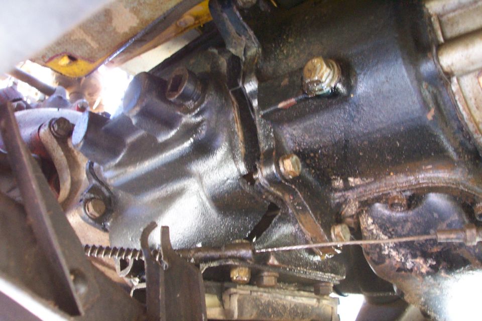 Close-up of black transmission and transfer case underside with linkage, spring, cable, and rusty bolts on a Jeep.