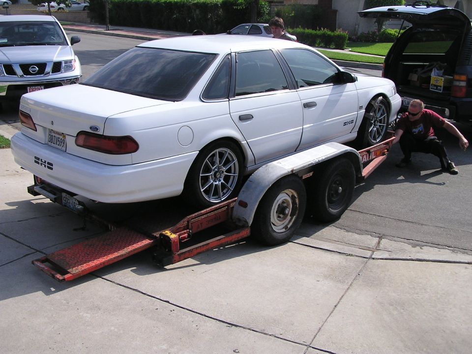 White 1993 Ford Taurus on a trailer, being loaded by a person. Another SUV is parked nearby