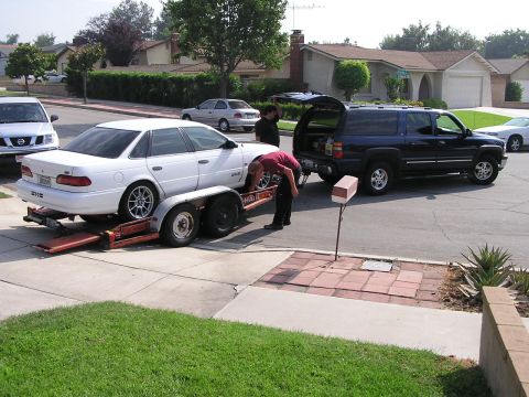 1993 Ford Taurus - A white 1993 Ford Taurus is loaded onto a trailer, with a dark SUV nearby.