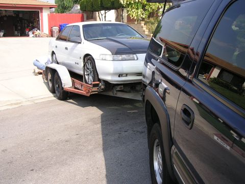 1993 Ford Taurus - A white 1993 Ford Taurus is loaded onto a trailer, being towed by a dark SUV.