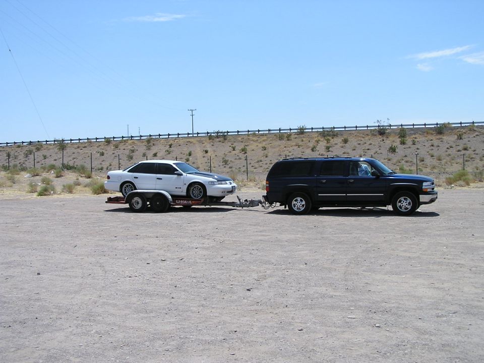 White sedan on a trailer, towed by a dark SUV, parked in a lot
