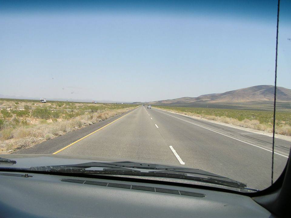 View from inside a vehicle looking out at a long, straight highway stretching into the distance under a clear blue sky