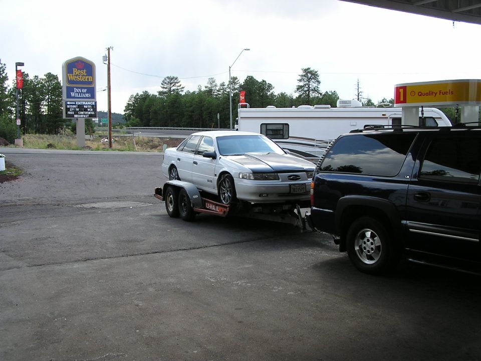1993 Ford Taurus on a trailer, towed by a dark SUV, near a Best Western and a gas station