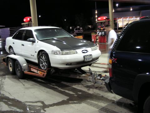 1993 Ford Taurus - A white sedan with a black hood is loaded onto a trailer at a gas station, with a dark SUV visible.
