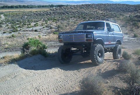 1986 Ford Bronco - A lifted Ford Bronco navigates a sandy, off-road trail in a dry, hilly landscape.