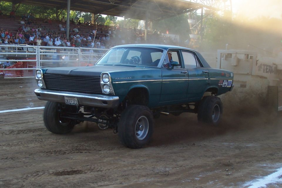 Teal sedan on huge tires, looks like it's in a mud pit or pulling event. Has 'Fairlane' badge on fender