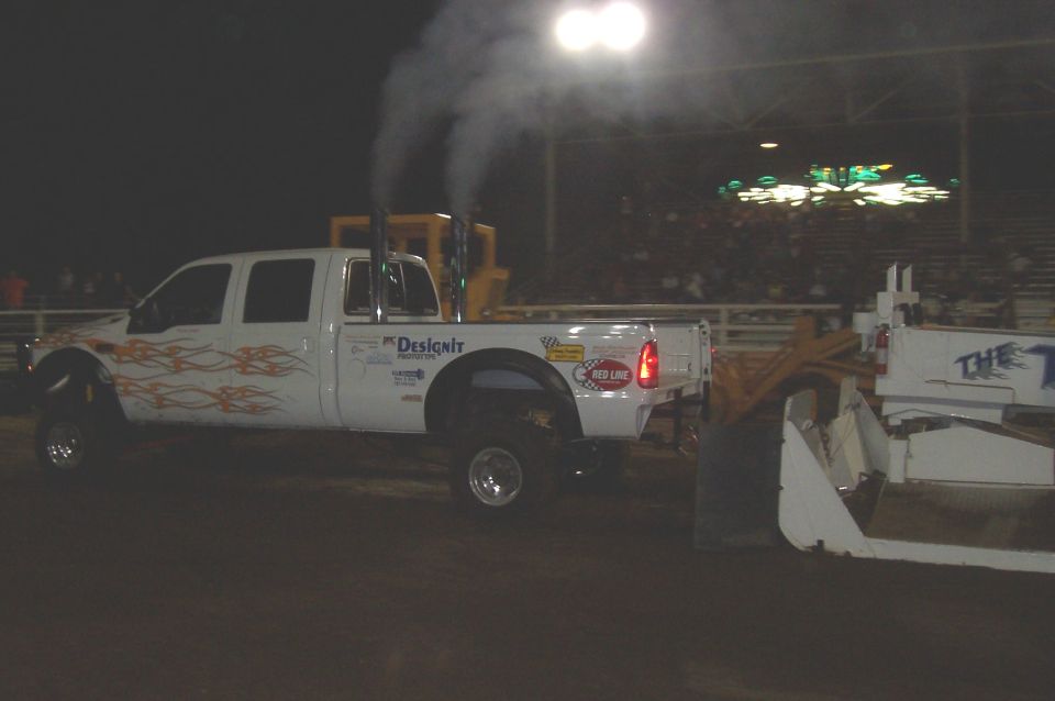 White pickup truck with flame decals, dual exhaust stacks, and "Designit" text on the side, pulling a large white implement. Crowd in background
