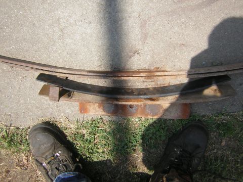 1995 Ford Bronco - Overhead shot of a multi-leaf spring pack laid on a concrete driveway with a rusty spring perch or shackle plate beneath it, photographed s…