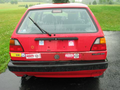 1991 Volkswagen Golf - Rear view of a red Volkswagen Golf GL hatchback covered in stickers, parked outdoors on a wet day.