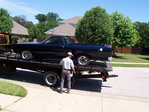1969 Ford Ranchero - A black pickup truck is being towed on a flatbed truck, with a man standing on the side of the road.