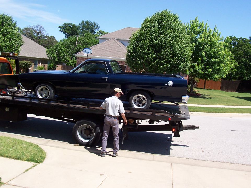 Dark Ford Ranchero on a flatbed tow truck in a suburban street, man securing it