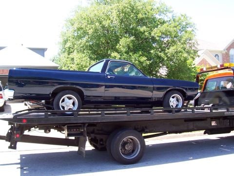 1969 Ford Ranchero - A dark blue Ford Ranchero is being towed on a flatbed truck, with a tow truck driver visible in the cab.