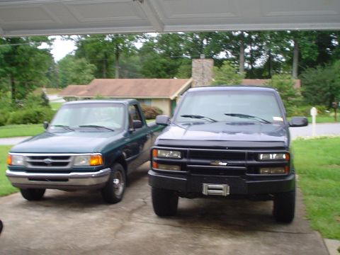 1981 Ford Bronco - Two pickup trucks are parked side-by-side in a driveway under a garage overhang.