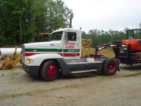 1981 Ford Bronco - A white semi-truck with distinctive red and green stripes is seen parked, hauling a mini-excavator on a trailer.