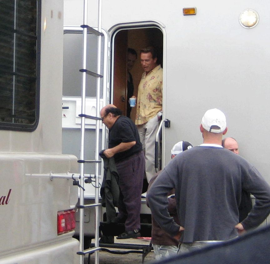 People around a white trailer doorway; a man in a floral shirt holds a cup inside