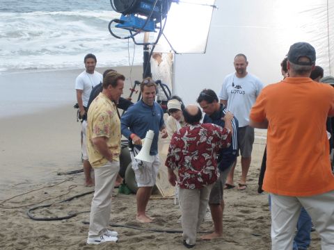 1990 Ford Bronco - film crew with a large lamp and reflector works on a beach around several people, including one in a floral shirt.