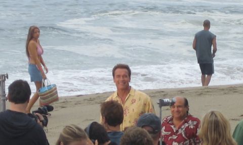 1990 Ford Bronco - man in a yellow floral shirt stands by the surf amid a film crew and crowd, with a bikini-clad woman walking past.