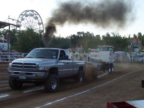 1994 Ford F-150 - Dodge Ram truck competes in a tractor pull, emitting heavy black smoke. A Ferris wheel is visible in the background.