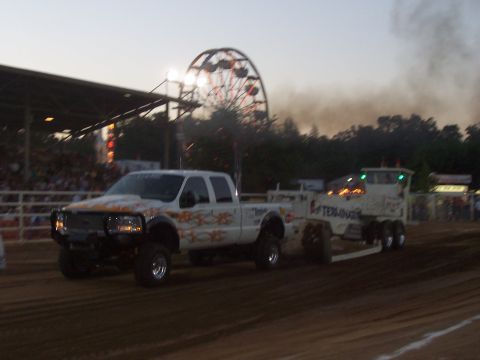 1994 Ford F-150 - A white pickup truck with flame decals tows a trailer past a Ferris wheel and grandstands at an outdoor event.