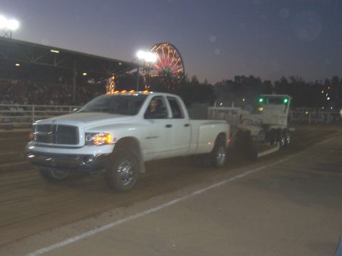 1994 Ford F-150 - A white Dodge Ram dually truck tows a trailer at a fairground event, with a Ferris wheel illuminated in the twilight sky.